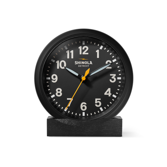 The Shinola 6" Runwell Clock features a black analog face with white numerals, white hour and minute hands, and a yellow second hand. It sits on an oak stand, highlighting Shinola craftsmanship against a white background.