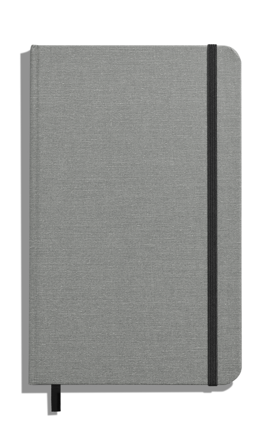The Shinola Medium Hard Ruled Journal - Stone, a closed gray fabric-covered linen journal with acid-free paper, features a black elastic band and ribbon bookmark, shown on a white background.