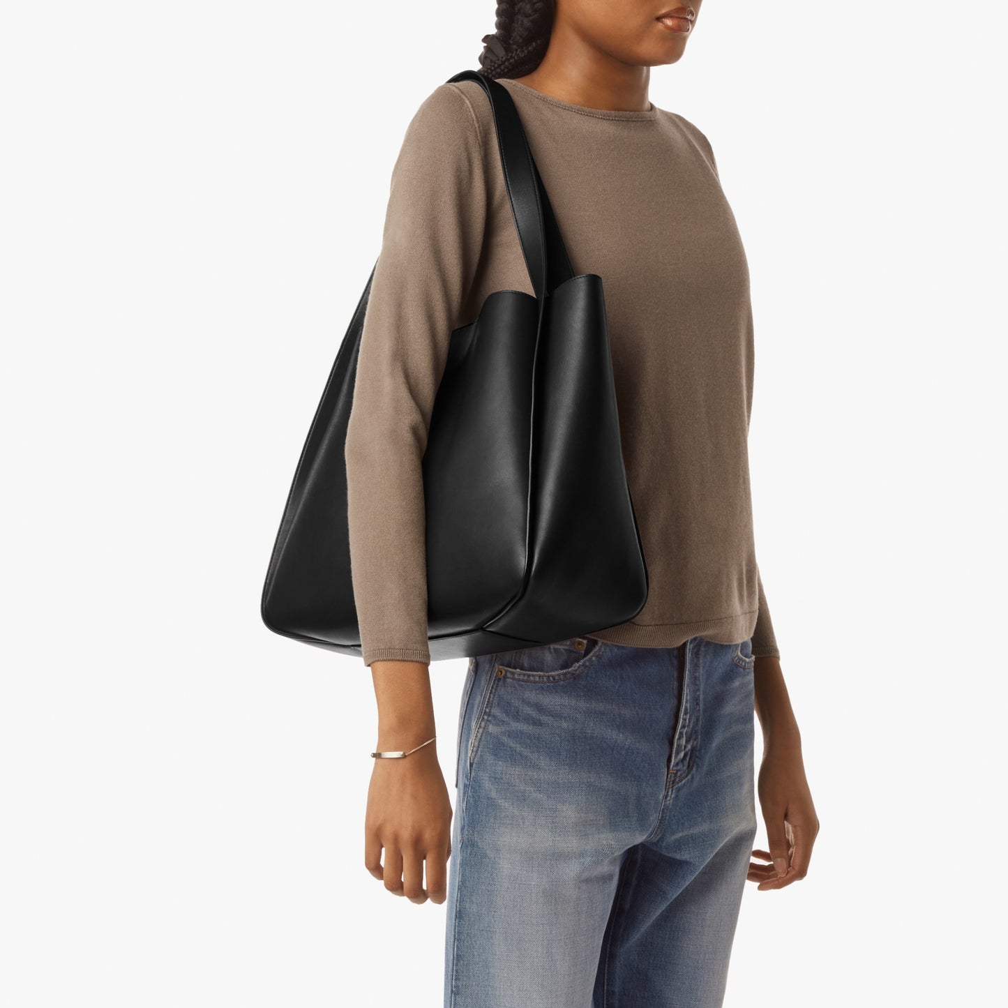 A woman in a taupe long-sleeve shirt and blue jeans carries the Shinola Derby Tote Bag - Black, made from natural leather, on her right shoulder against a plain white background.