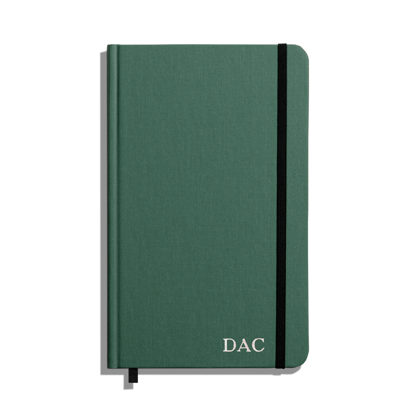 The Shinola Medium Hard Ruled Journal - Fern is a green linen hardcover notebook with a black elastic band, acid-free paper, ribbon bookmark, and white DAC initials embossed on the front cover’s bottom right corner.