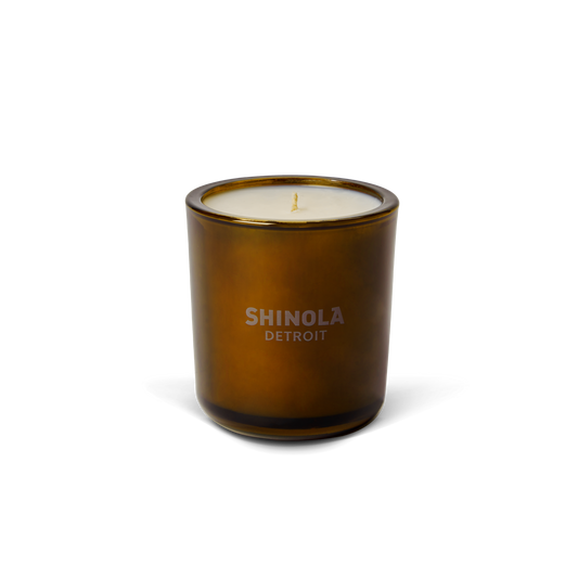 A brown glass container holds the Shinola Smoke 8oz Scented Candle, made with white coconut soy wax and a woody scent that captures Detroit fall, shown against a plain white background.