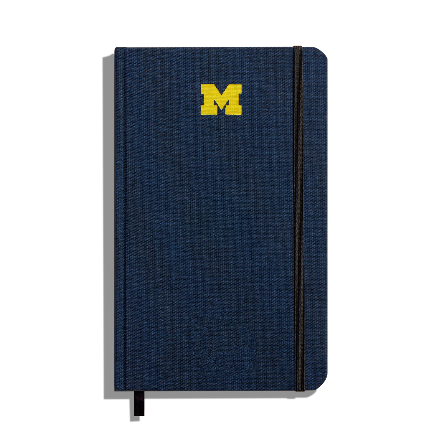 The Shinola Medium Hard Ruled Journal in navy features a yellow University of Michigan M logo on the cover, a black elastic band closure on the right side, and a black ribbon bookmark at the bottom.