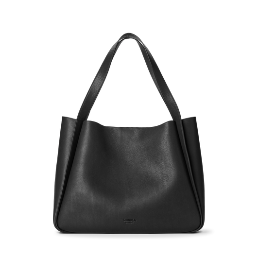 The Shinola Derby Tote Bag - Black is crafted from natural leather with wide, flat shoulder straps and a smooth, minimalist design. This soft-structured everyday bag features subtle branding near the bottom center.