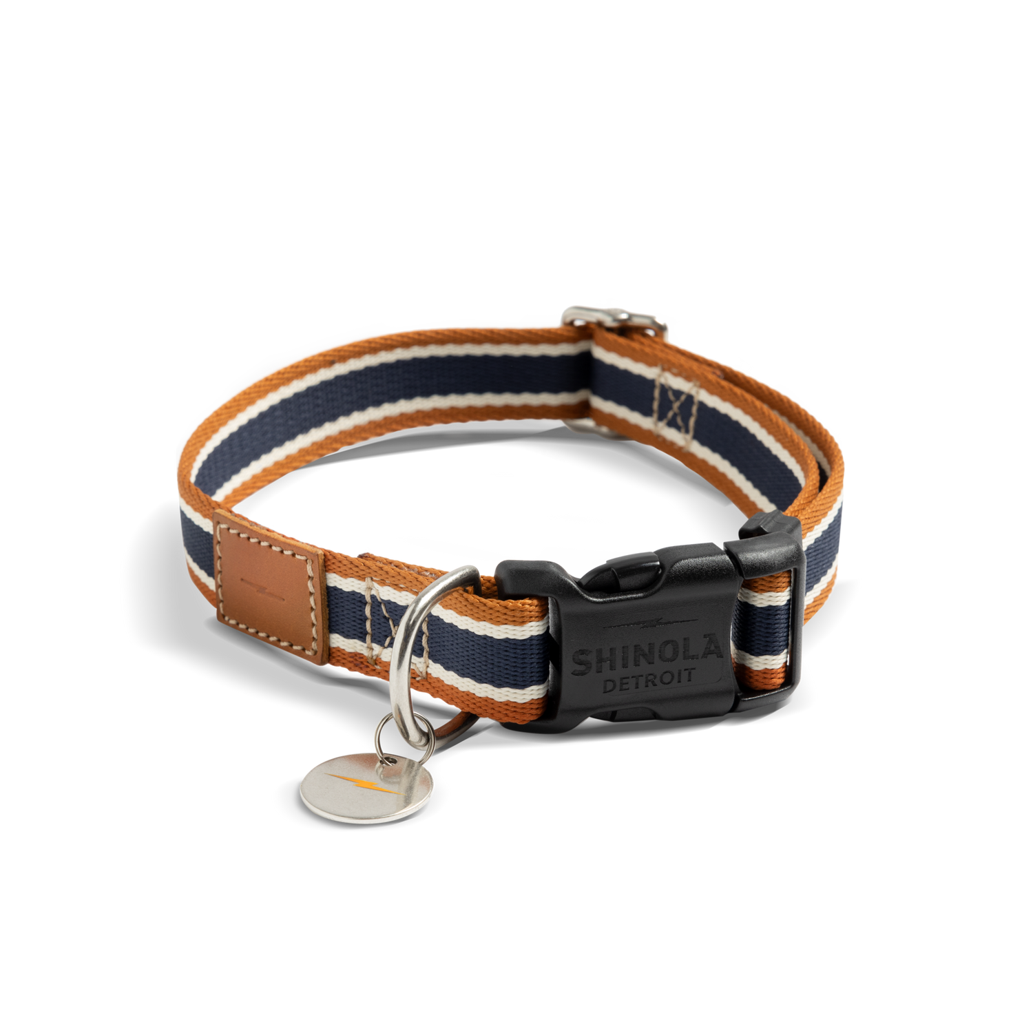 The Shinola Nylon Dog Collar - Brand Stripe features orange, navy, and white stripes, a black Shinola Detroit buckle, silver ring, brown leather patch accent, and a round silver tag.