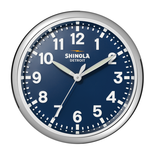 A 14" Shinola Runwell Clock in navy, featuring a round design with a blue face, white numbers, and silver frame. The clock displays 10:10:34 with "Shinola Detroit" below the 12 o'clock mark.