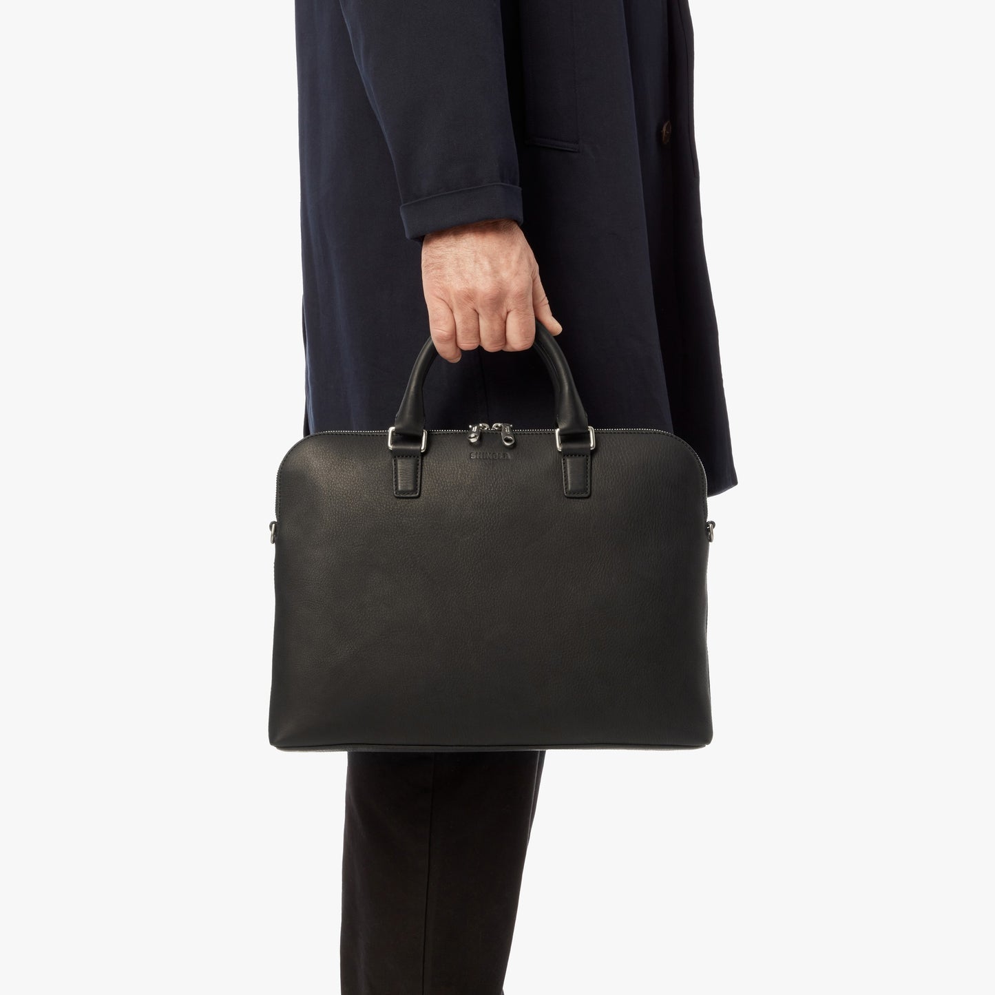 A person in a dark coat and pants holds the Shinola Portfolio Briefcase - Black by its handles, displayed against a plain white background.