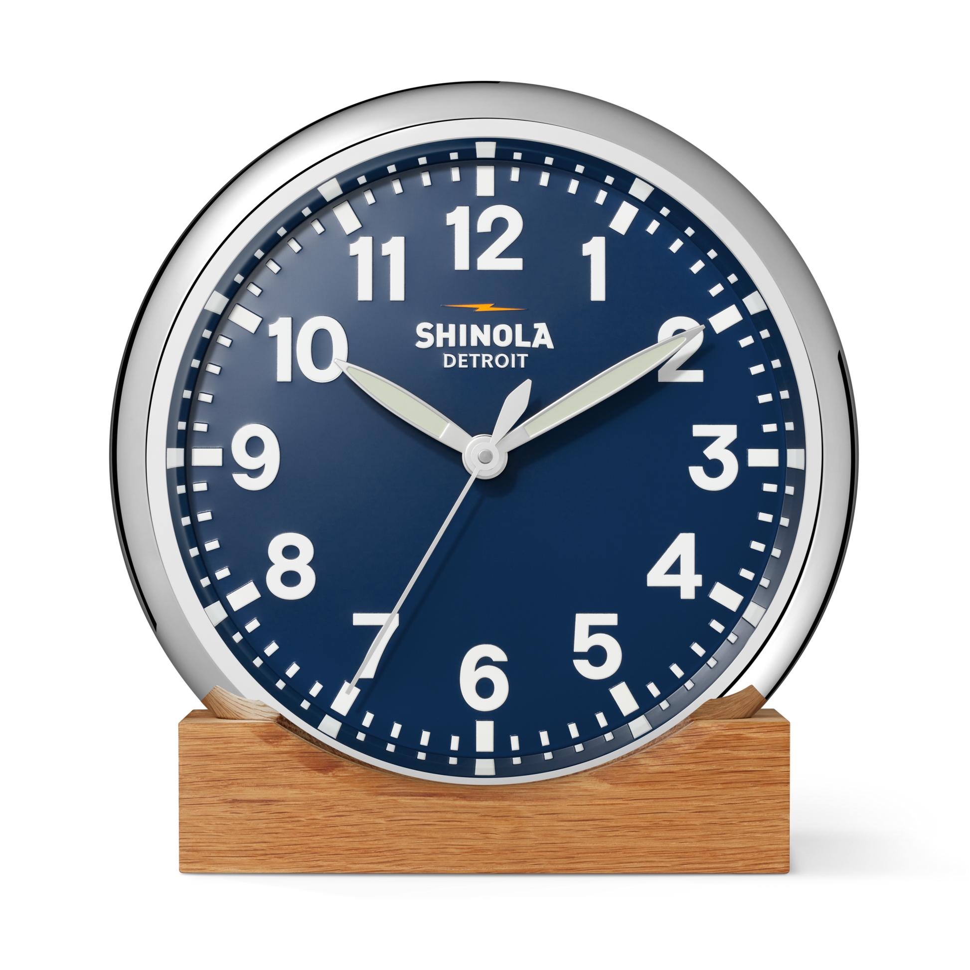 A Shinola 14" Runwell Clock in navy features a round blue face with white numbers and hands, set in a chrome frame atop a wooden base. The time displayed is 1:51:34 against a plain white background.