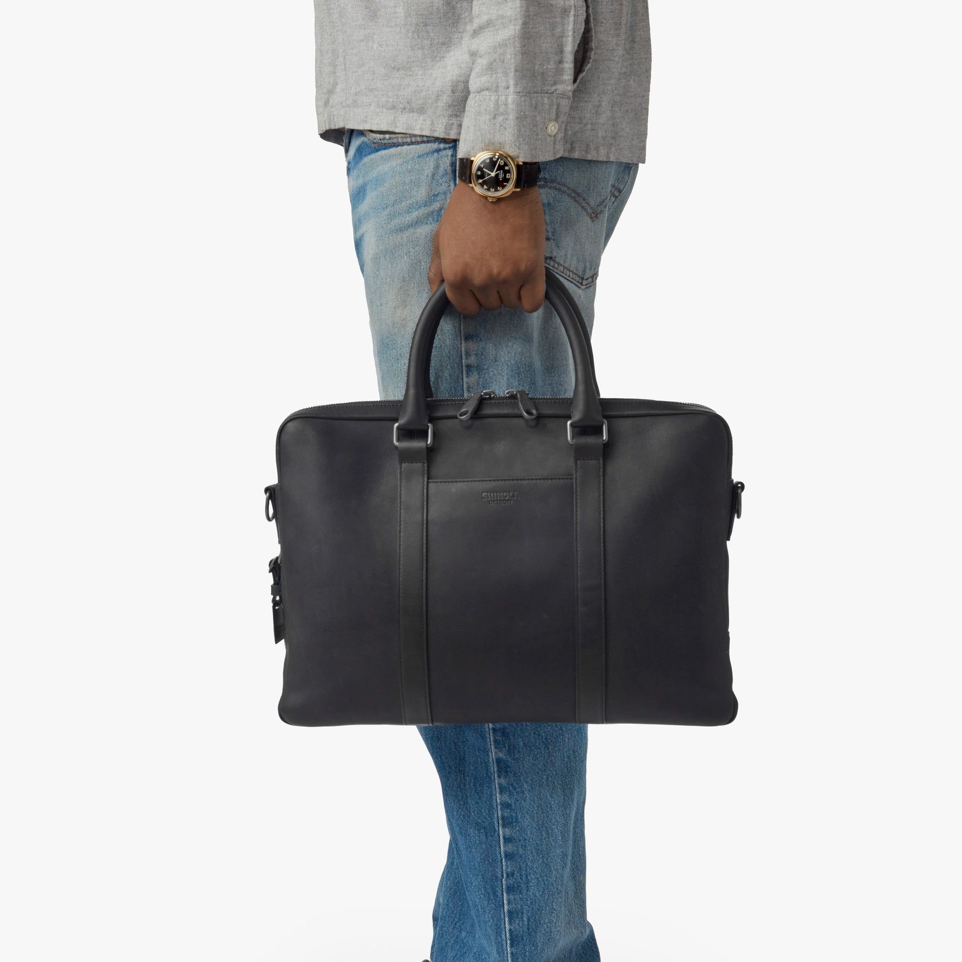A person in a gray shirt, blue jeans, and a watch holds the nicelaundry-jp.computer Briefcase - Black by its handles against a plain white background.