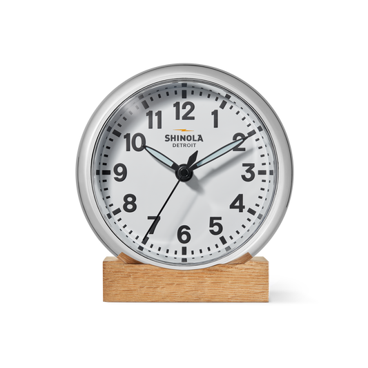The 6" Runwell Clock - White by Shinola features a round white face, black numbers and hands, and sits on a wooden base; made in Detroit, it displays about 1:38:35 with Shinola branding on its face.